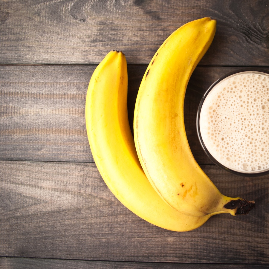 Glass of banana smoothie and fresh bananas on brown wooden background. Top view
